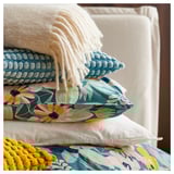 Cama com almofadas florais coloridas, incluindo azul-petróleo, laranja e branco. Uma manta de malha branca e um cobertor texturizado amarelo também são visíveis.