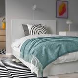 Quarto moderno com cama branca, manta cinza-turquesa, tapete listrado, arte, luminária. O cobertor é de poliéster reciclado e elegante.