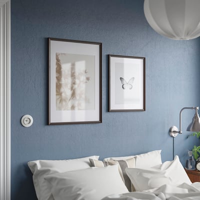 Relógio analógico acima da cama, branco com números pretos. Dois quadros: abstrato, borboleta. Cama com travesseiros brancos, paredes azuis.