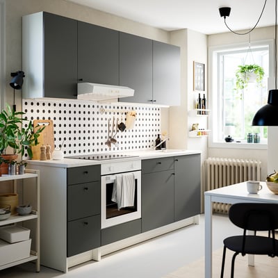 Cozinha moderna com armários cinzentos, balcão em mármore, backsplash de bolinhas, forno, cadeiras pretas, plantas.