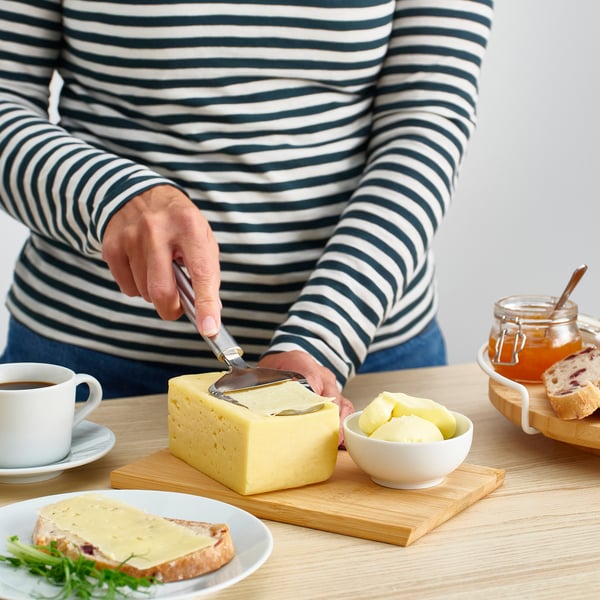 Uma pessoa fatia queijo em uma tábua de madeira usando um cortador de aço inoxidável. Café, queijo no pão e maçãs também estão presentes.
