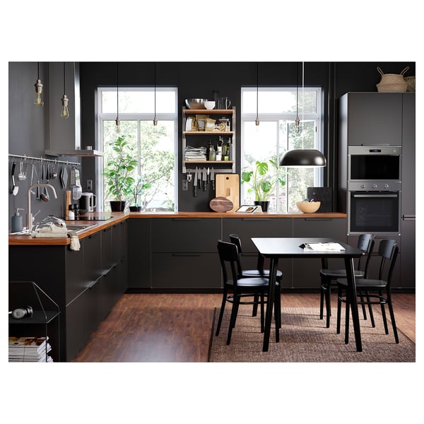 Cozinha moderna com armários escuros, balcão de madeira, eletrodomésticos de metal, cadeiras pretas, luzes e plantas.