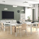 Sala de conferências moderna com mesas brancas, cadeiras creme, paredes verdes, duas tvs. Arranjo elegante, piso de madeira.