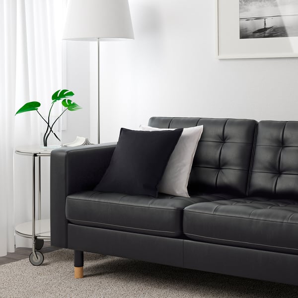 Sala de estar moderna com sofá de couro preto, almofadas de destaque, mesa lateral, abajur, planta e foto emoldurada na parede.