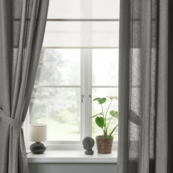 Janela com cortinas cinza amarradas para trás, mostrando vista do lado de fora. O peitoril branco segura lâmpada, planta, decoração. A luz é filtrada.