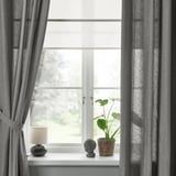 Janela com cortinas cinza amarradas para trás, mostrando vista do lado de fora. O peitoril branco segura lâmpada, planta, decoração. A luz é filtrada.
