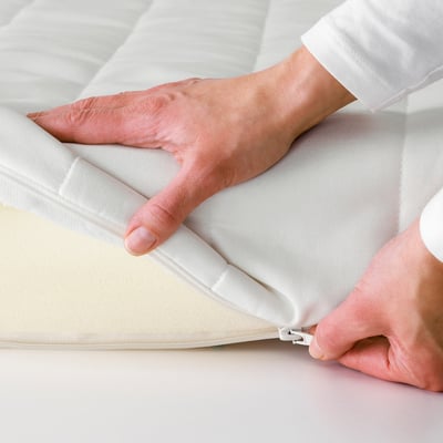 Mãos fechando um colchão branco, mostrando sua capa removível para facilitar a limpeza.