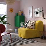 Sala de estar brilhante com sofá de cadeira amarela, decorada com almofada de ponto preto, prateleira verde próxima e poltrona vermelha, em tapete listrado preto-branco.