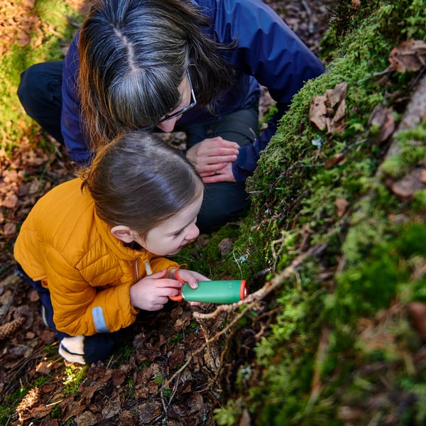 Uma criança usa uma lanterna led com manivela na floresta. Um adulto observa, enfatizando o aprendizado e a exploração.