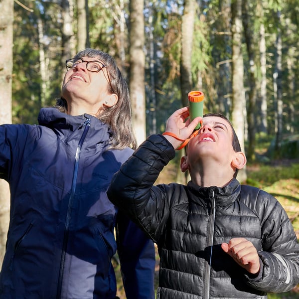 Duas pessoas em uma floresta, uma segura uma criança usando um pequeno telescópio verde e laranja, olhando para cima.