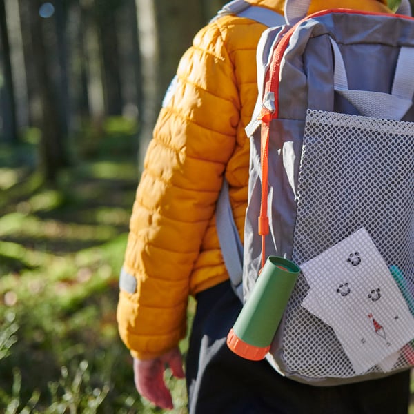 Criança vestindo uma jaqueta laranja carrega uma mochila com uma garrafa de água e um bloco de notas em um ambiente ao ar livre.