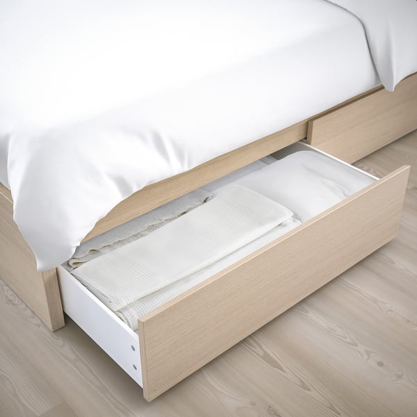 Cama com gaveta aberta, mostrando espaço de armazenamento. Madeira clara, lençóis brancos.
