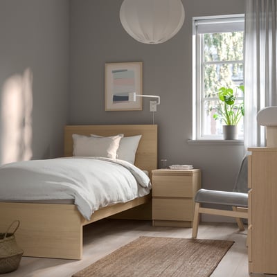 Quarto minimalista: paredes cinza, móveis de madeira clara. Cama MALM com lençóis brancos, plantas e livros na mesa de cabeceira. Janela grande.