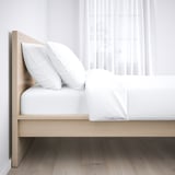 Cama minimalista moderna com cabeceira de madeira clara, lençóis brancos e piso escuro.