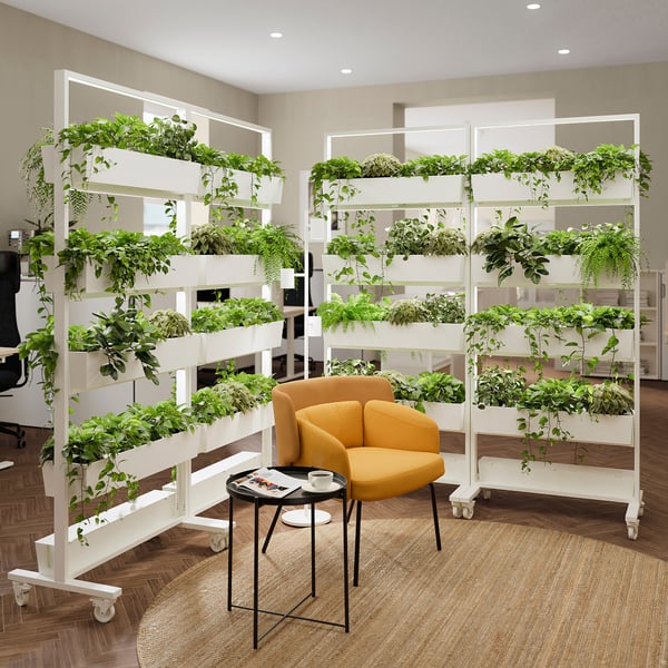 A parede verde modular MITTZON divide a sala com estruturas e rodas altas de plantas, ao lado de cadeira laranja e mesa lateral preta, adicionando uma atualização vibrante.