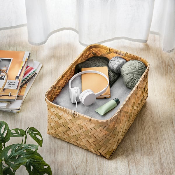 Cesta de bambu tecida contendo fones de ouvido, fios e um livro em um piso de madeira, ao lado de uma pilha de livros e um vaso de plantas.