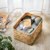 Cesta de bambu tecida contendo fones de ouvido, fios e um livro em um piso de madeira, ao lado de uma pilha de livros e um vaso de plantas.