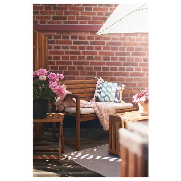 Pátio externo com móveis de madeira, parede de tijolos, flores rosa, almofadas e um tapete.