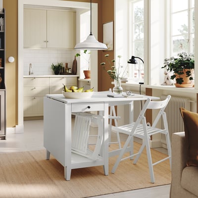 Pequeno conjunto de jantar branco, com cadeiras dobráveis e uma mesa de folhas rebatíveis com armazenamento, bem guardado em uma cozinha aconchegante.