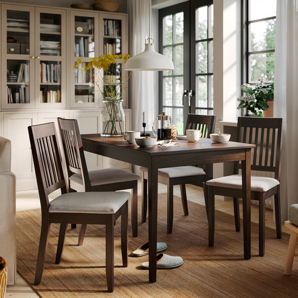 Área de jantar aconchegante com mesa NÄSINGE, cadeiras cinza, flores, estante e janelas amplas.
