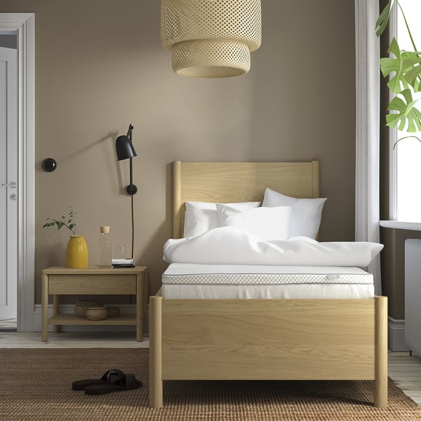Quarto minimalista com cama de madeira clara, roupa de cama branca, mesa de cabeceira com decoração, luminária pendente, janela grande e tapete.