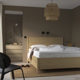 Quarto mínimo com móveis de madeira, roupa de cama branca e um espelho redondo. A cama é focal, maior que a cadeira preta, sob a lâmpada pendente.