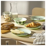 Conjunto de loiça OFTAST verde sobre mesa de madeira. Inclui pratos com croissants, queijo, pão e utensílios.