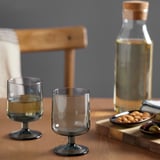 Dois copos transparentes sobre uma mesa de madeira, um cheio de líquido. Ao lado, uma garrafa de vidro com uma rolha de madeira e uma tigela de madeira cheia de petiscos.