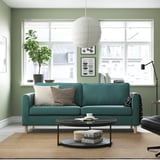 Sala de estar moderna com sofá azul-petróleo de 3 lugares, paredes verdes, janelas grandes e pernas de madeira. Almofadas completam o espaço.