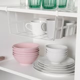 Um armário de cozinha com prateleiras mostrando uma cesta de armazenamento com clipe segurando canecas, acima de tigelas e pratos rosa e branco empilhados.