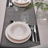 Mesa elegante com pratos e tigelas brancas, talheres, copos em jogos americanos estampados.