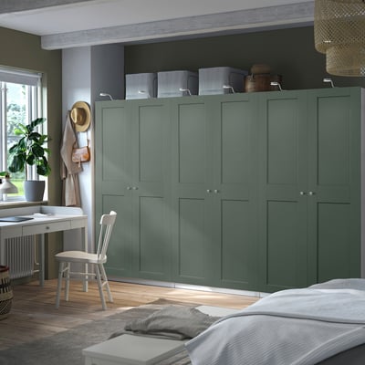 O grande guarda-roupa verde PAX fica alto perto de uma mesa branca, com chapéus pendurados e caixas de armazenamento em cima.