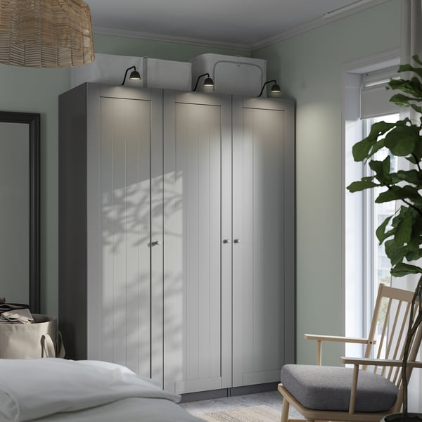 Quarto minimalista com guarda-roupa PAX cinza claro, alto e largo, cama combinando, cadeira pequena e planta grande. Iluminação suave.