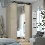 Quarto minimalista com guarda-roupa PAX bege, centro espelhado, cadeira branca, cama, roupas penduradas, arte e planta.