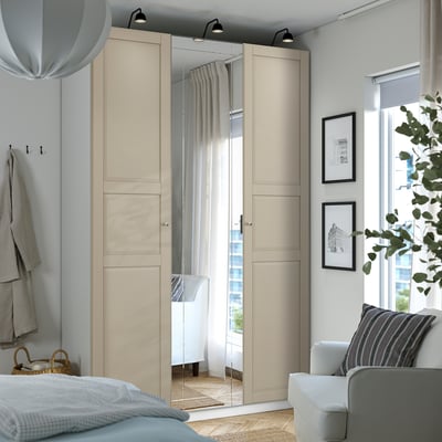 Roupeiro PAX moderno com portas bege, espelhos, dobradiças de fecho suave. Nas proximidades, poltrona branca, luminária e janela com cortinas transparentes.