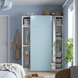 Um quarto moderno com um guarda-roupa PAX azul claro com portas de correr revelando roupas organizadas, prateleiras e cestos de armazenamento.