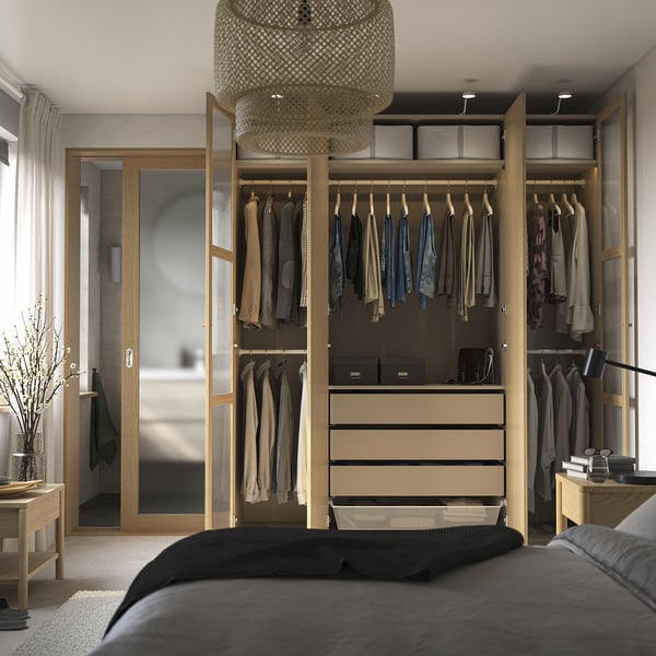 Quarto com guarda-roupa moderno aberto mostrando roupas, gavetas e prateleiras. Luminária e sala conectada visíveis.