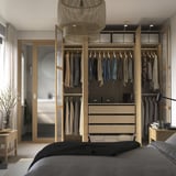 Quarto com guarda-roupa moderno aberto mostrando roupas, gavetas e prateleiras. Luminária e sala conectada visíveis.