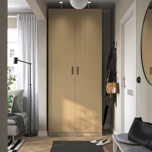 Roupeiro PAX em madeira entre porta e janela, arrumação de roupa e acessórios, com acabamento em madeira clara em sala minimalista.