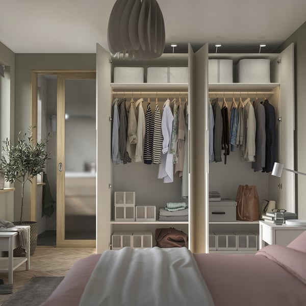 Quarto moderno com guarda-roupa PAX aberto exibindo roupas, bolsas e caixas de armazenamento. Uma cama aconchegante com lençóis rosa está em primeiro plano.