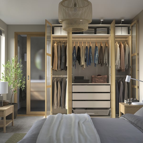 Armário moderno de madeira com portas de vidro, iluminação e cama com lençóis cinza/branco.