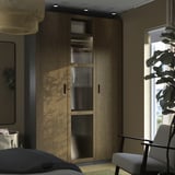 Quarto moderno com guarda-roupa alto de madeira PAX, cadeira cinza, planta e janela.