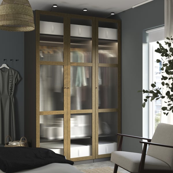 Quarto moderno com guarda-roupa alto PAX em carvalho, com portas de vidro e dobradiças de fechamento suave, perto de uma cadeira aconchegante e janela grande.