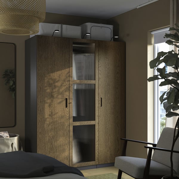 Um quarto moderno com um roupeiro alto PAX em madeira, portas com fecho suave e caixas de arrumação cinzentas. Uma janela e uma planta adicionam luz natural e vegetação.