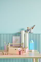 Área de estudo com parede azul, mesa de madeira, caixa de arte, copos coloridos, lâmpada lápis, desenho emoldurado.