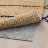 Um tapete tecido bege com detalhes em amarelo e azul está sobre um piso de madeira, mostrando sua parte inferior com forro de feltro cinza para evitar escorregões.