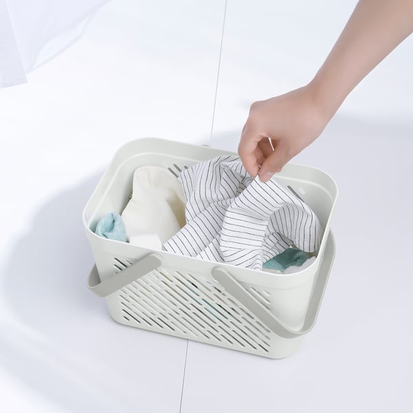 A mão coloca o item em uma cesta de armazenamento RÅNEN branca cheia de toalhas e produtos de higiene pessoal, mostrando seu uso prático para organizar itens essenciais do banheiro.