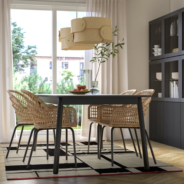 Sala de jantar moderna com mesa preta, cadeiras de vime, tapete bege e preto, luminária de madeira, janela grande.