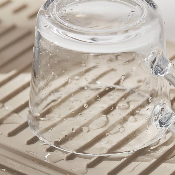Copo de vidro transparente com gotas de água fica em um tapete texturizado bege, provavelmente um tapete de secagem expansível e aderente.
