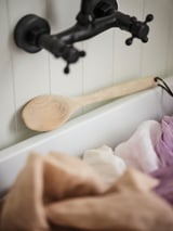 Utensílio de cozinha de madeira: espátula pendurada perto de torneira preta, pia branca, sabão e toalha.